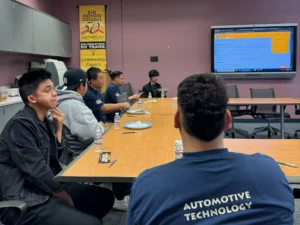Group of students sitting around a table in a classroom, focusing on a presentation displayed on a screen, with one wearing an "automotive technology" shirt.