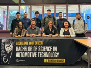 Group of ten people smiling behind a banner reading "bachelor of science in automotive technology at rio hondo college" in a classroom setting.
