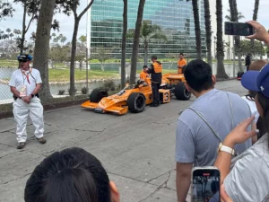 A vintage orange race car with the number 3, surrounded by spectators and race staff, parked on a tree-lined street with office buildings in the background.