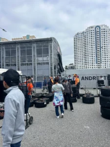 People and workers walking near a large racing event setup with stacks of tires, a viewing structure, and city buildings in the background.