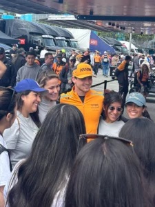 A man in an orange cap and shirt smiling and posing for a photo with six women in a busy outdoor setting.