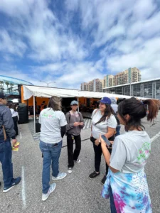 Group of people conversing at an outdoor event with tents and a food truck, wearing casual clothing and some with branded t-shirts.