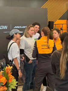 Group of people, including two team members in mclaren uniforms, engaged in a conversation in a motorsport team hospitality area.
