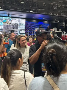 A man speaks into a microphone addressing a crowd in an indoor entertainment venue with a racing-themed backdrop.