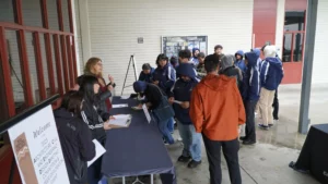 A group of people, some in hooded jackets, are gathered outside near a registration table under a covered area on a rainy day. A woman at the table is assisting attendees.