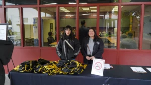 Two women stand behind a table with black and yellow drawstring bags and a "No Smoking" sign, in front of a building with large windows.