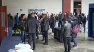 A group of people in jackets gathers outside a building on a wet day, some standing in line near tables with informational materials.
