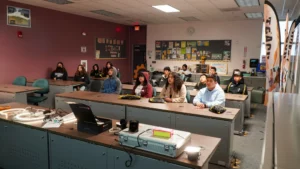 A group of people sit attentively in a classroom with desks facing forward. Various educational materials are visible on the walls, and a projector setup is on the instructor's desk at the front.