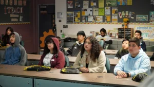 A group of people, some wearing hoodies and jackets, sit attentively in a classroom. Various posters and notices are on the walls, and a monitor displays information behind them.