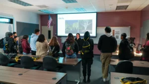 A group of students stand in a classroom facing a speaker. A screen behind the speaker displays an image and the word "theia." The room has desks, chairs, and an American flag.