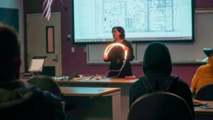 A person stands at the front of a classroom holding a U-shaped light, with diagrams on a large screen behind them. Several students sit and watch attentively.