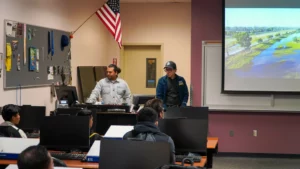 Two men stand at the front of a classroom, speaking to students. An American flag is displayed on the wall, and a screen shows an outdoor scene. Computer stations are set up in front of the students.