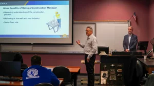 Two men stand at the front of a classroom during a presentation. A slide displayed on the projector screen reads "Other Benefits of Being a Construction Manager." Students sit facing the presenters.