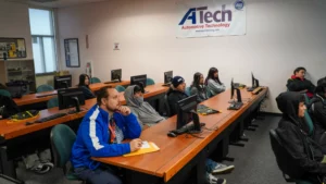 A group of people are seated in a classroom with computers, listening attentively to a presentation at ATech Automotive Technology.
