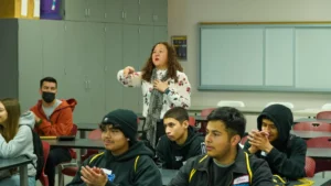A woman is speaking to a group of students in a classroom. Some students are wearing hoodies and beanies, and a few are clapping. A man in a mask is seated on the side.