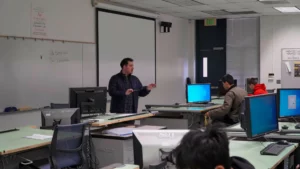 A man stands at the front of a classroom, gesturing while speaking. Several students sit at desks with computers, listening. A large projection screen is behind the speaker.