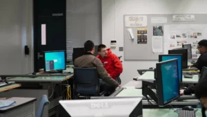 Two men sit and converse in a computer lab filled with various workstations. The room has overhead lighting, a bulletin board with posted papers, and multiple monitors displaying different screens.