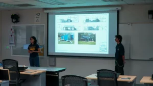 Two people stand at the front of a classroom presenting architectural designs projected on a screen. Tables with chairs fill the room, and a sign on the wall says "NO food or drink.
