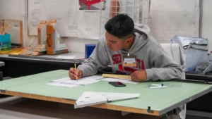 A man wearing a grey hoodie and ID badge sits at a drafting table, working on architectural plans with a pencil and ruler.