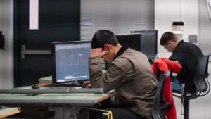Two individuals are working on computer-aided design (CAD) software at desks in a computer lab. The person in the foreground is focusing on the screen.