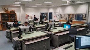 A computer lab with several people working at desktop computers, desks arranged in rows, and a few individuals standing or moving in the background.