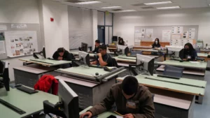Several students work individually at drafting and computer workstations in a classroom setting, focusing on their projects.