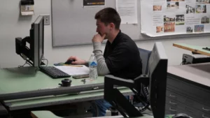 A person is sitting at a desk, focused on a computer screen, with a hand on their chin, a water bottle, papers, and a phone nearby. Another computer monitor and a bulletin board are in the background.