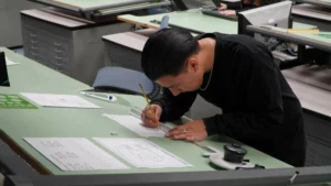 A person in a black shirt is leaning over a green drafting table, focused on drawing precise lines with a pencil on a technical drawing. Various drafting tools and documents are scattered on the table.