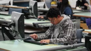 A student wearing glasses and a plaid shirt works on a computer in a classroom with other students.
