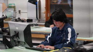 A person in a blue jacket is focused on typing at a desktop computer in a classroom or office setting. A second person is in the background, also working.