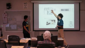 Two individuals are standing in front of a projection screen in a classroom, presenting architectural plans. One person is pointing at the screen, and a seated audience is observing.