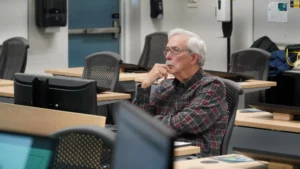 An older man with gray hair and glasses, wearing a plaid shirt, is sitting in a classroom with computer monitors, attentively listening.