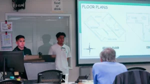 Two men stand beside a screen displaying architectural floor plans during a presentation. An audience member sits at a table observing. A sign indicating "No food or drink" is visible on the wall.