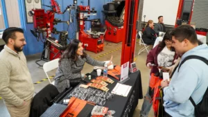 People interact at a booth with pamphlets on a table, in a room with red machinery and tools in the background.