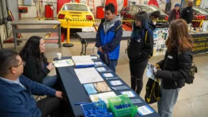 People gather around a table displaying pamphlets and documents at an information booth in a workshop with vehicles in the background.