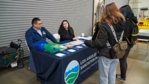 Two individuals sit behind a table with promotional materials labeled "Los Angeles Sanitation District," engaging with three people standing in front of them in a warehouse setting.