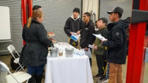 A group of people stands around a table with informational materials and small buckets in what appears to be an indoor event or workshop setting.