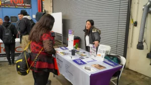 A woman in a red and black outfit speaks to another woman standing behind a booth with informational pamphlets at a career fair or expo. A corrugated metal door is closed in the background.