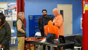 Three men are standing and discussing near a table with drones and a laptop in a workshop setting. One man is holding an orange drone case. The background includes a blue wall, tools, and workshop equipment.