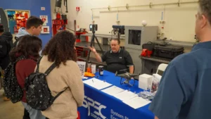 A man in a black polo shirt discusses equipment at a booth with four attendees in front of him. The booth is covered with pamphlets and small tech devices, with industrial machinery in the background.