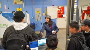 A woman in a suit and hat shakes hands with a person wearing a backpack in an industrial setting. Several other people are gathered around, and a laptop is visible on a table nearby.