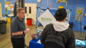 A man in a dark polo shirt stands near a display booth with a "Bentley: Advancing Infrastructure" banner, talking to a person in a black beanie and white hoodie. The booth has a bowl of candies.