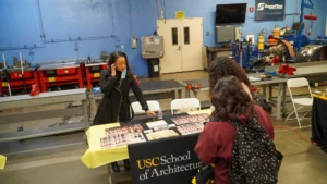 A woman stands behind a table displaying brochures and materials with a sign saying "USC School of Architecture" as three other people look at the display in a workshop setting.