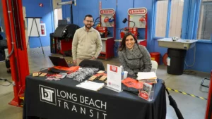 Two individuals at a Long Beach Transit booth, equipped with informational materials and promotional items, set up inside a workshop with machinery and safety equipment in the background.