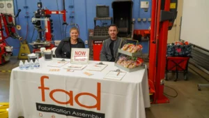 Two individuals sit at a booth with a "Now Hiring" sign, snacks, and water bottles, promoting "Fabrication Assembly" at a job fair in a workshop setting with tools and equipment in the background.