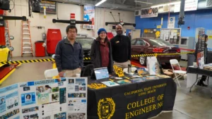 Three people stand behind a table covered with promotional materials for California State University Long Beach College of Engineering, set up in an automotive workshop. A project display board is on the left.