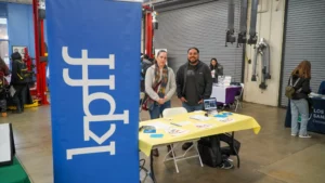 Two people stand behind a table with yellow tablecloth at an indoor event next to a blue banner displaying "kpfT." Other tables and attendees are in the background.