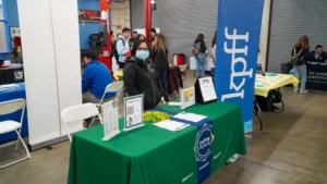 A person stands behind a green table with a KPFF banner at a career or information fair. The table has informational materials, including brochures and a QR code, and people are visible in the background.