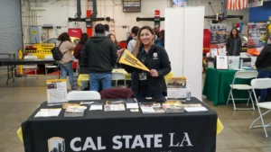 A representative at a Cal State LA booth holds a yellow pennant. The booth has flyers, brochures, and promotional materials. Other booths and people are in the background of the indoor space.