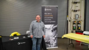 A man in a plaid shirt stands near a table with a Stantec banner that reads, "We are a community of scientists, planners, and engineers." The table has a black cloth and the area has yellow table covers.